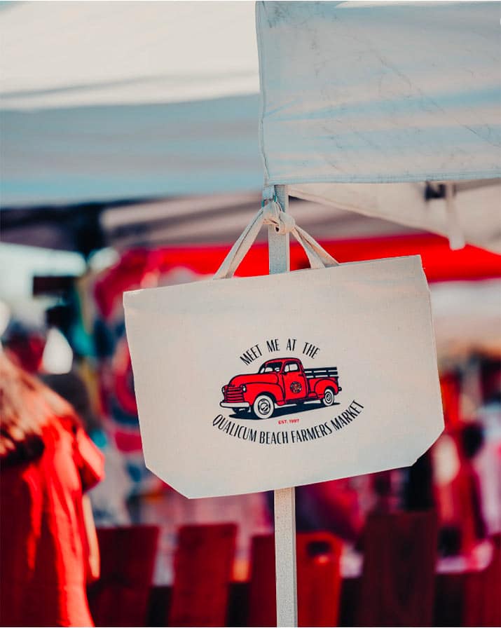 The Qualicum Beach Farmers Market logo on a canvas tote bag, hanging from a vendor stall at the Qualicum market.