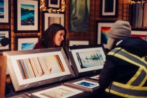 A female artist smiles behind her display of paintings, chatting to a customer at the Qualicum market.